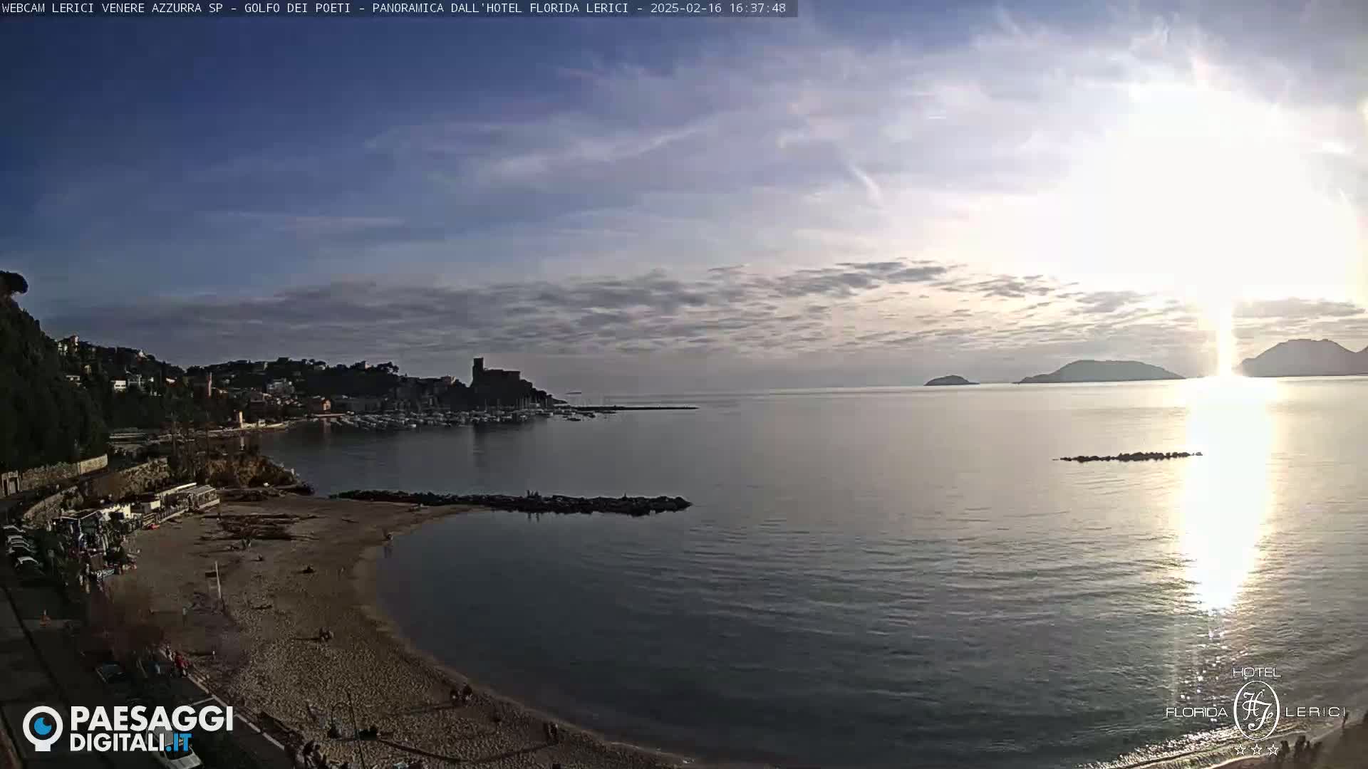 A sandy beach curves into a calm bay, with a town and distant hills visible under a partly cloudy sunset sky.