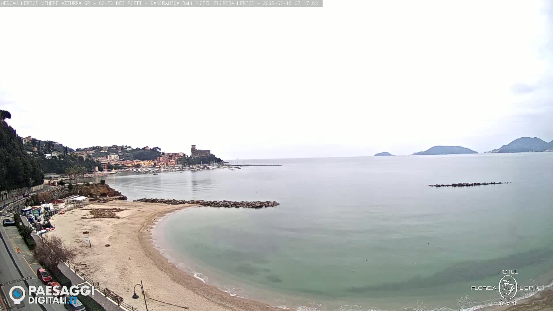 A sandy beach curves into a calm, light-green sea under an overcast sky, with a town and distant hills visible in the background.