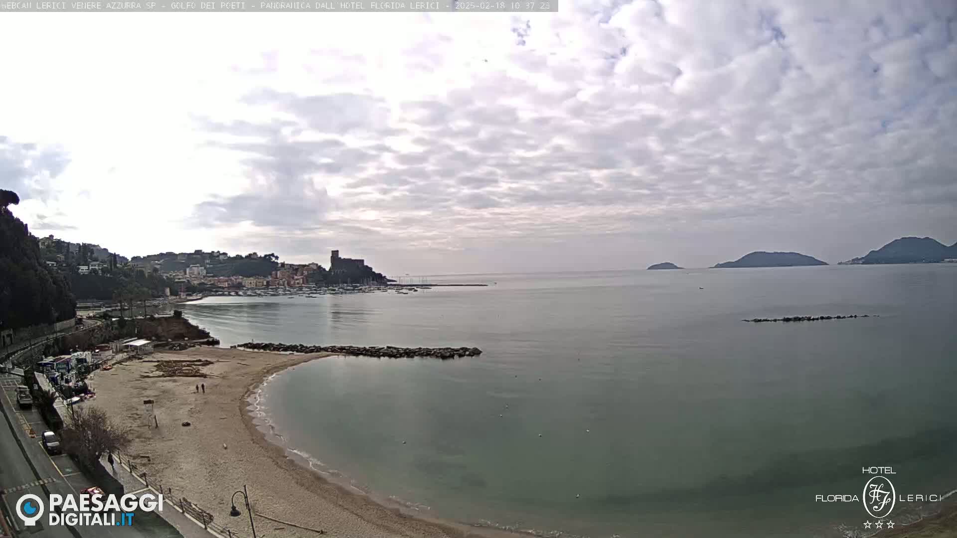 A sandy beach curves into a calm bay, with a town and hills visible in the distance under a cloudy sky.