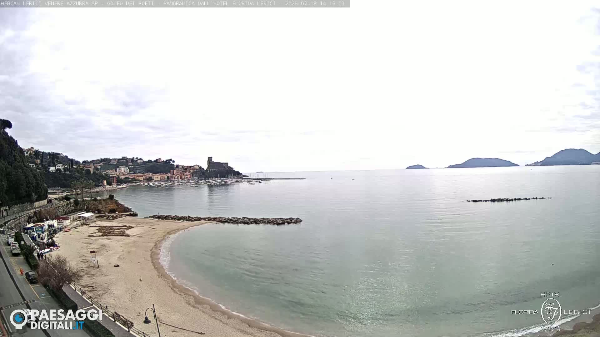 A sandy beach curves into a calm bay, with a town and distant hills visible across the water under an overcast sky.