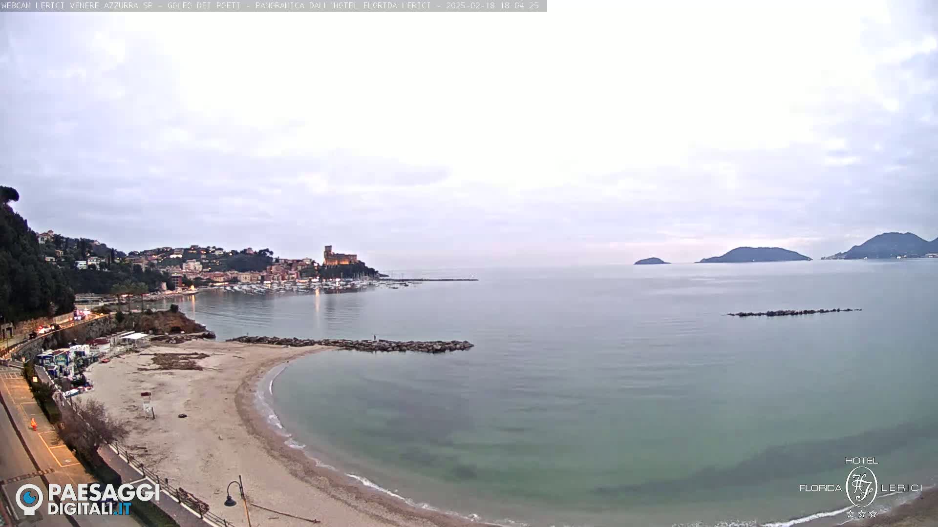 A tranquil coastal scene shows a sandy beach curving into calm, greenish water, with a town and a castle visible in the distance under an overcast sky.
