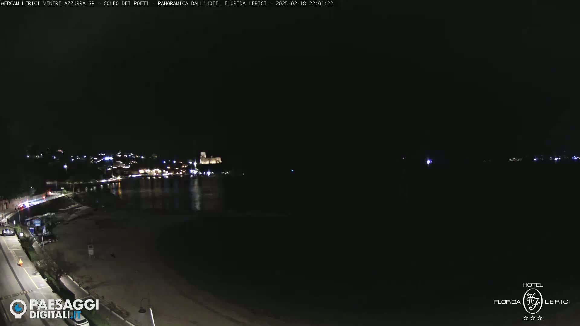 A dark, nighttime view of a beach and calm water with a town's lights visible in the distance.