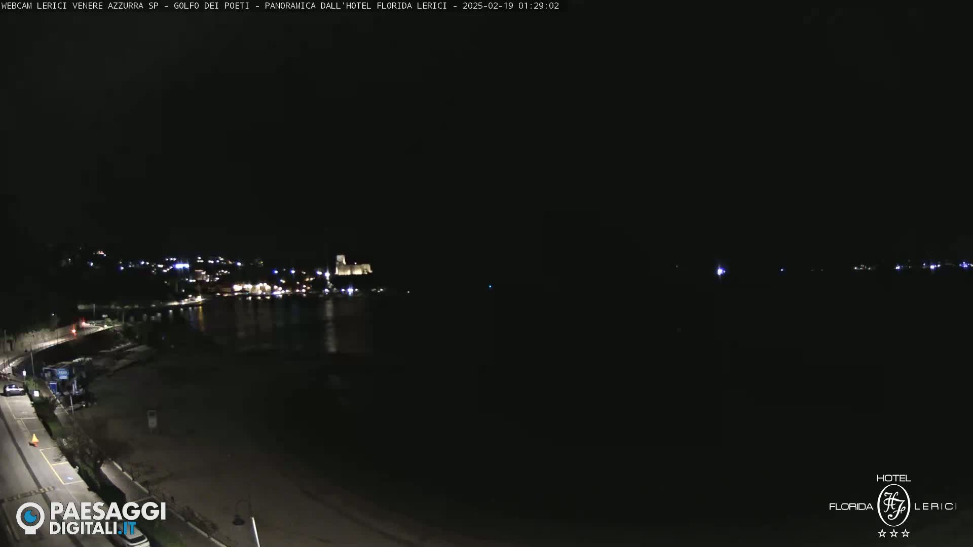 A nighttime view of a dark beach and a town across a body of water, with dimly lit buildings visible in the distance.