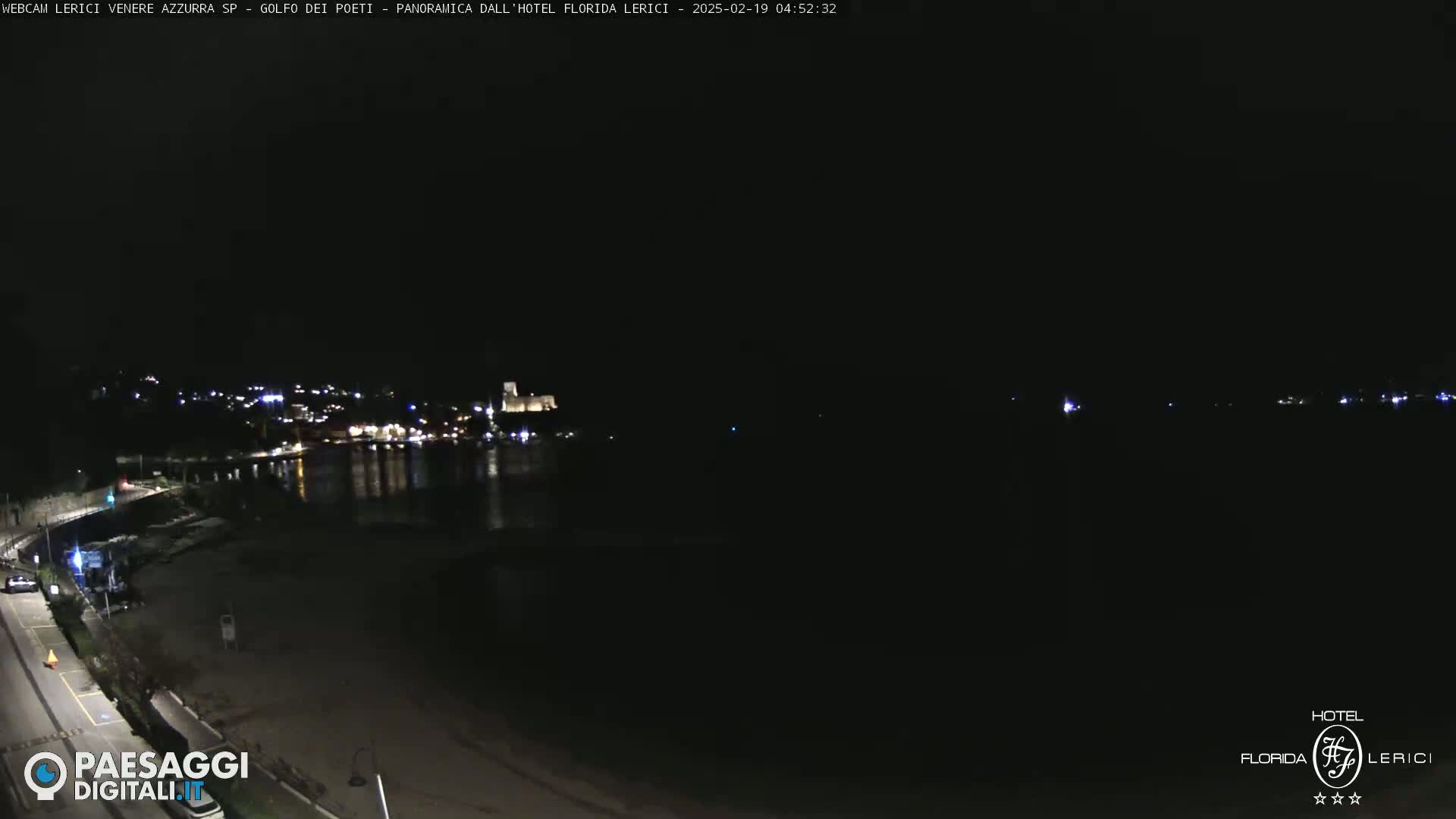 A nighttime long shot of a dark beach and calm water, with a town's lights visible across the water.