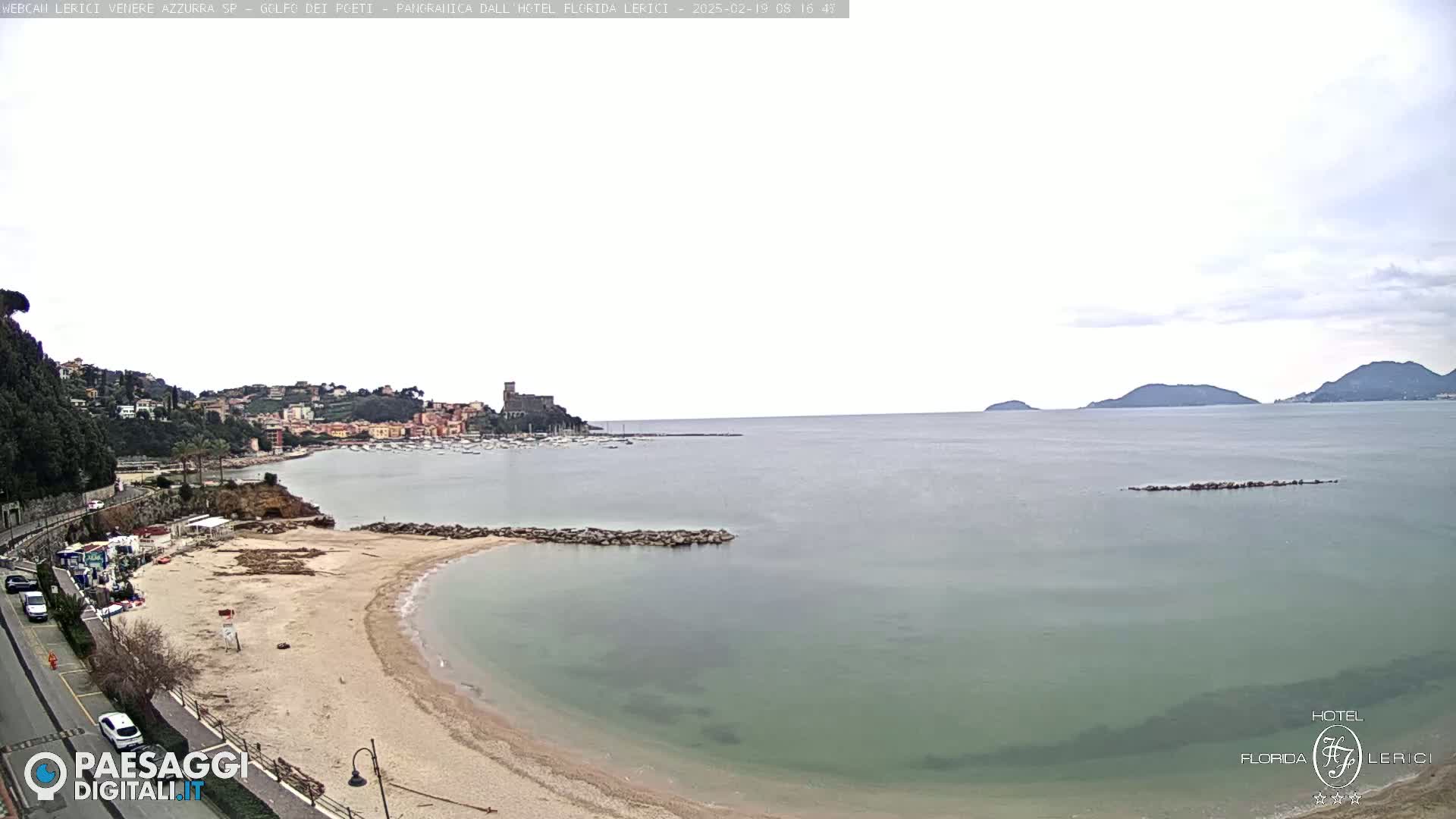 A sandy beach curves into a calm, pale green sea under an overcast sky, with a town and distant hills visible in the background.