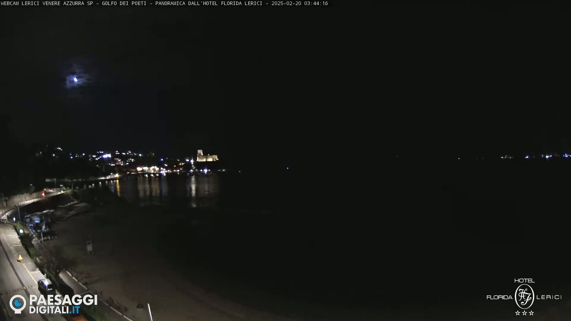 A nighttime coastal scene shows a dark beach and calm water reflecting a moonlit town in the distance.