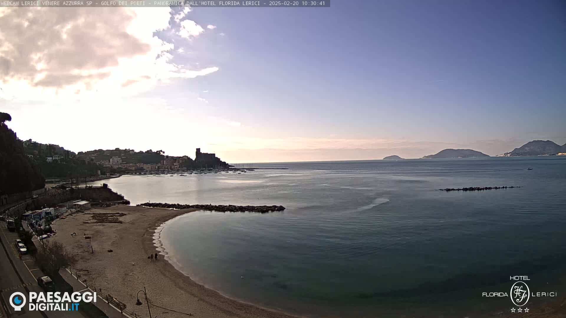 A mostly sunny day reveals a coastal town with a sandy beach and calm water, overlooking a bay and distant islands.