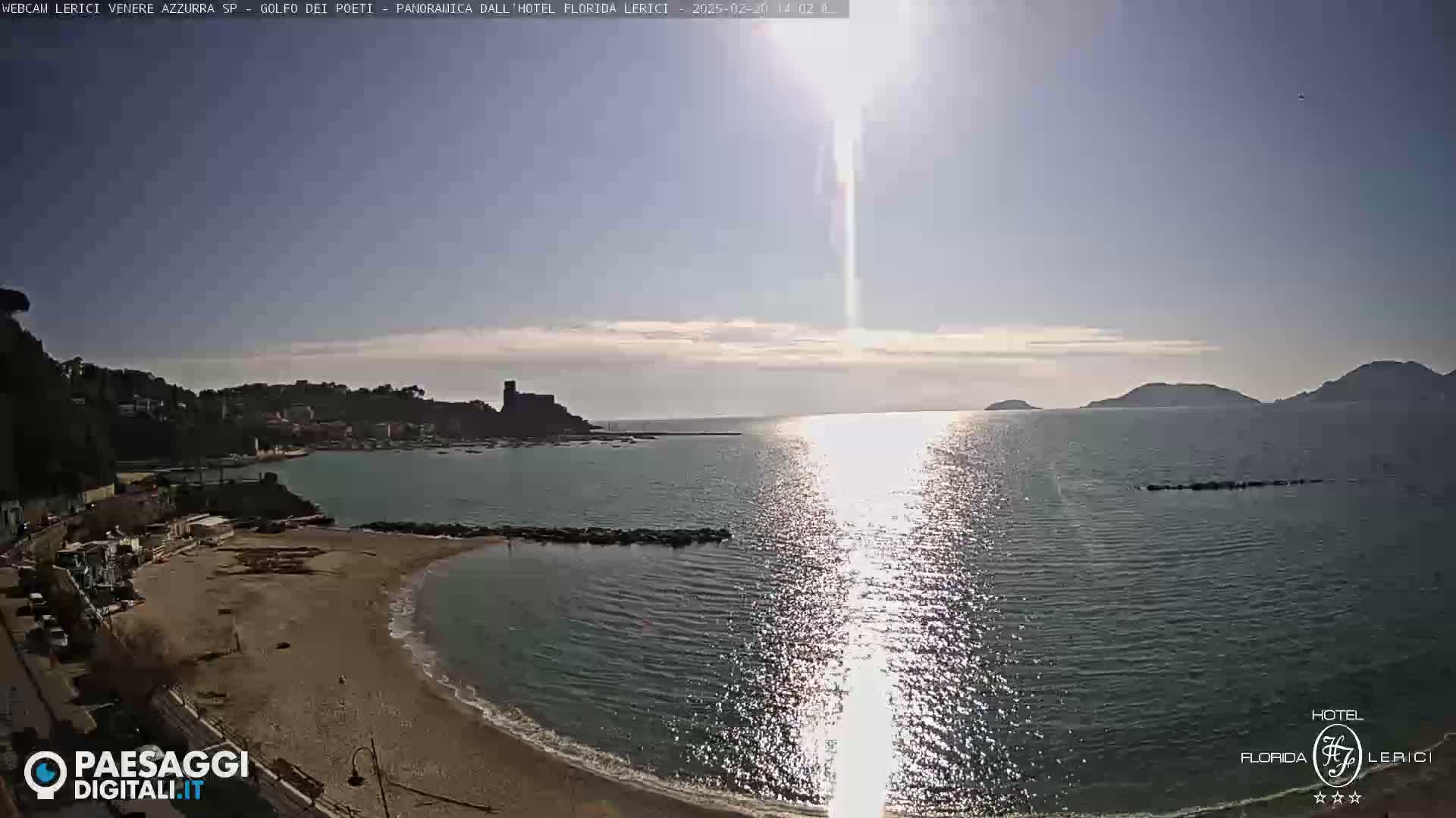 A sandy beach curves into a calm bay, with a town and distant hills visible under a bright, sunny sky.