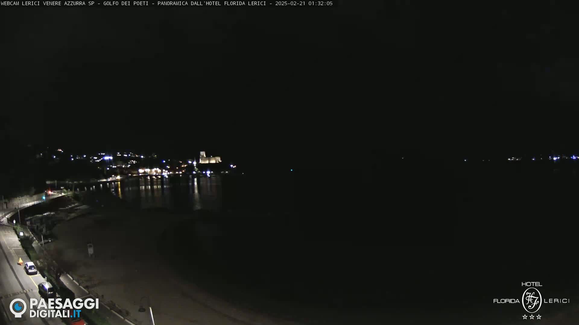 A nighttime, long shot of a dark beach and calm body of water with a town’s lights faintly visible across the water.