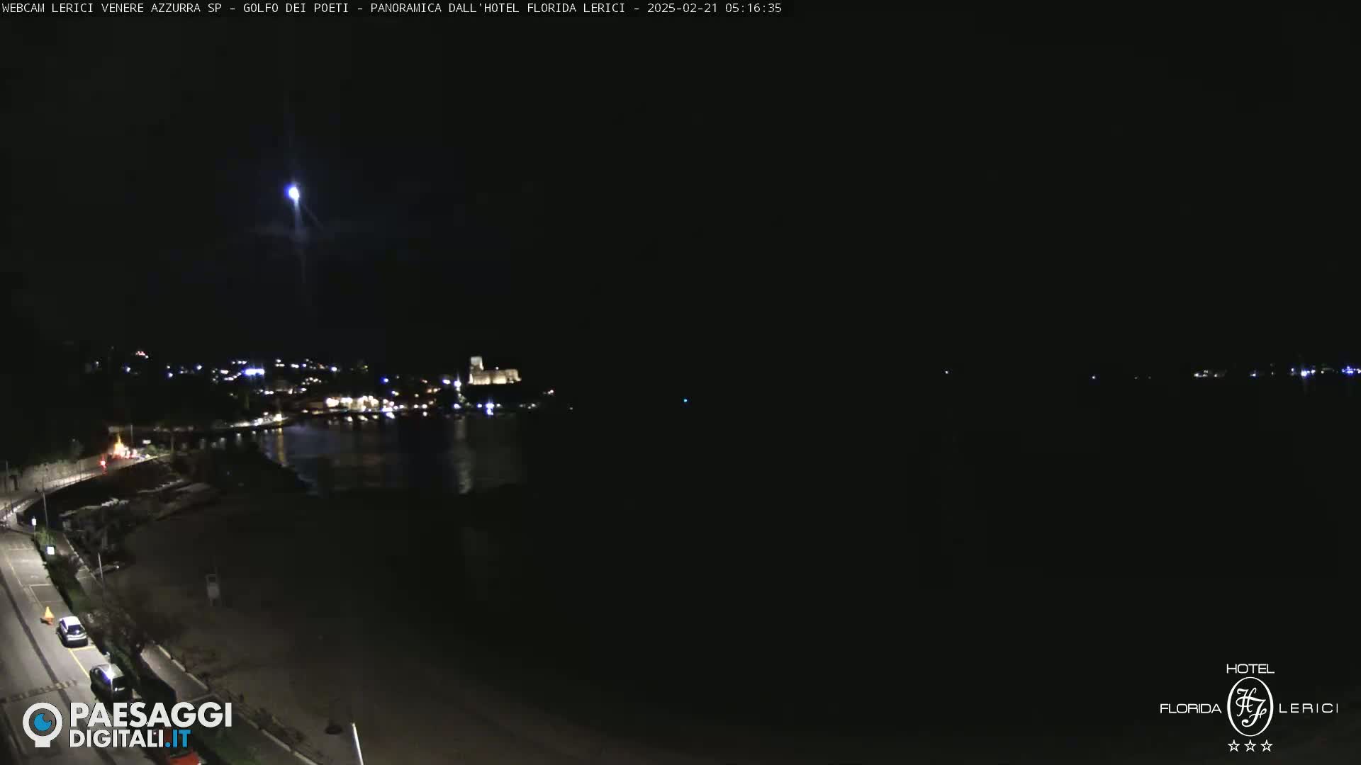 A nighttime long shot of a dark coastline with a town and a body of water, illuminated by scattered lights, and a bright light streak in the sky.