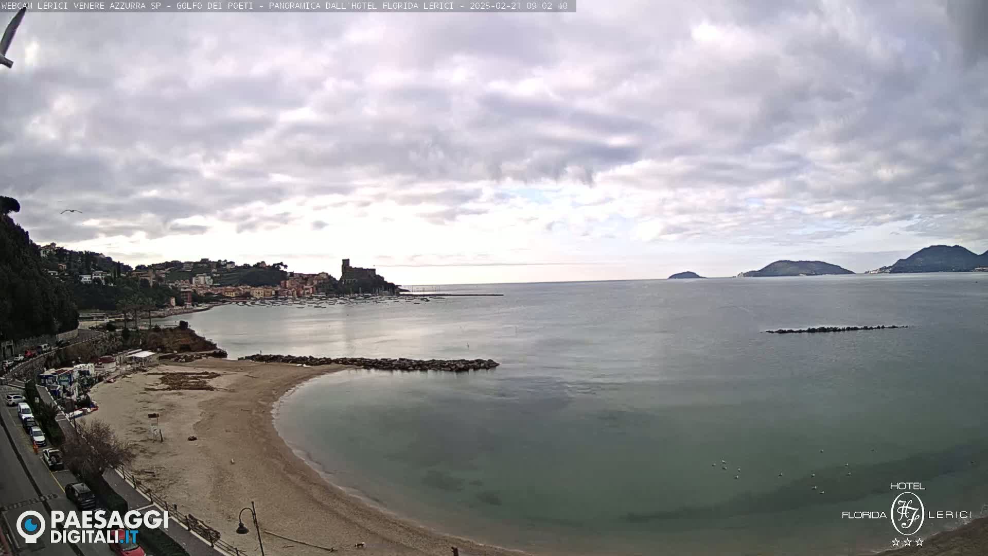 A mostly cloudy day reveals a tranquil bay with a sandy beach, a small town nestled on a hillside, and distant islands under a pale sky.