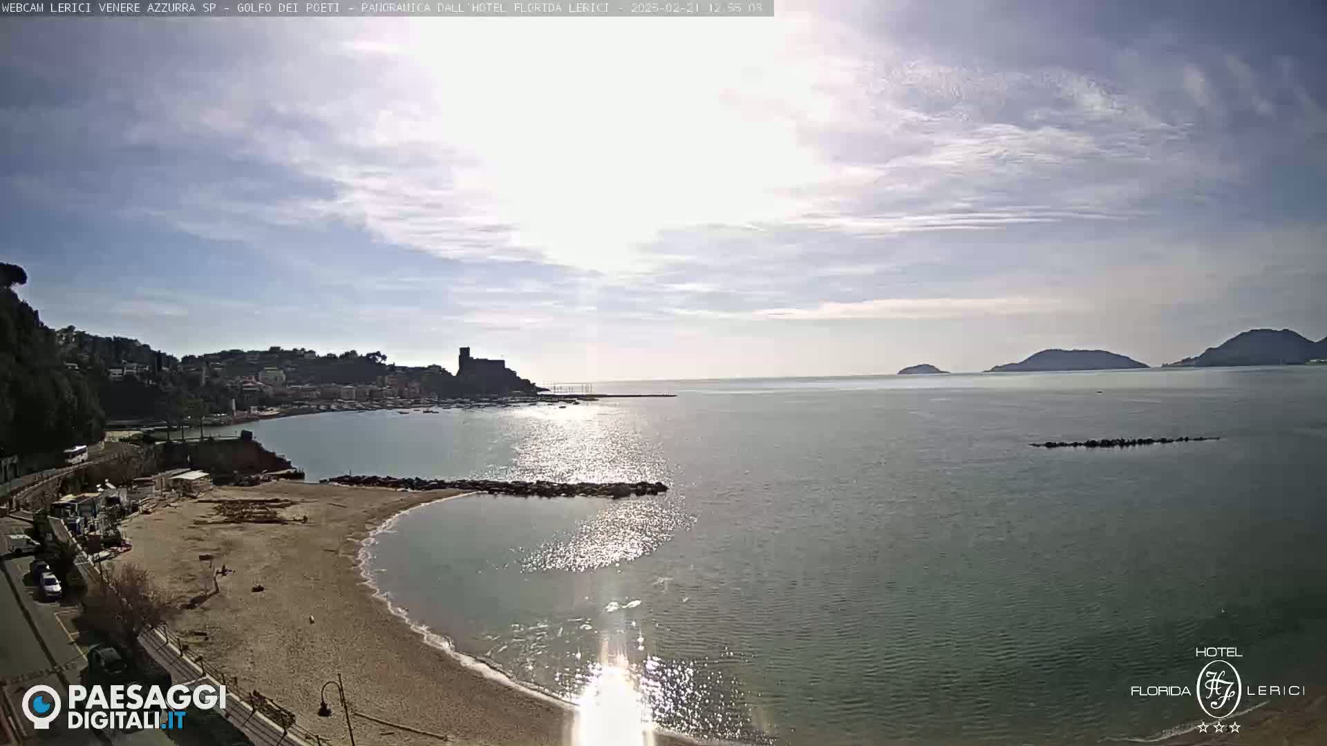A sunny day reveals a coastal town nestled beside a calm bay, with distant hills and a sandy beach in the foreground.