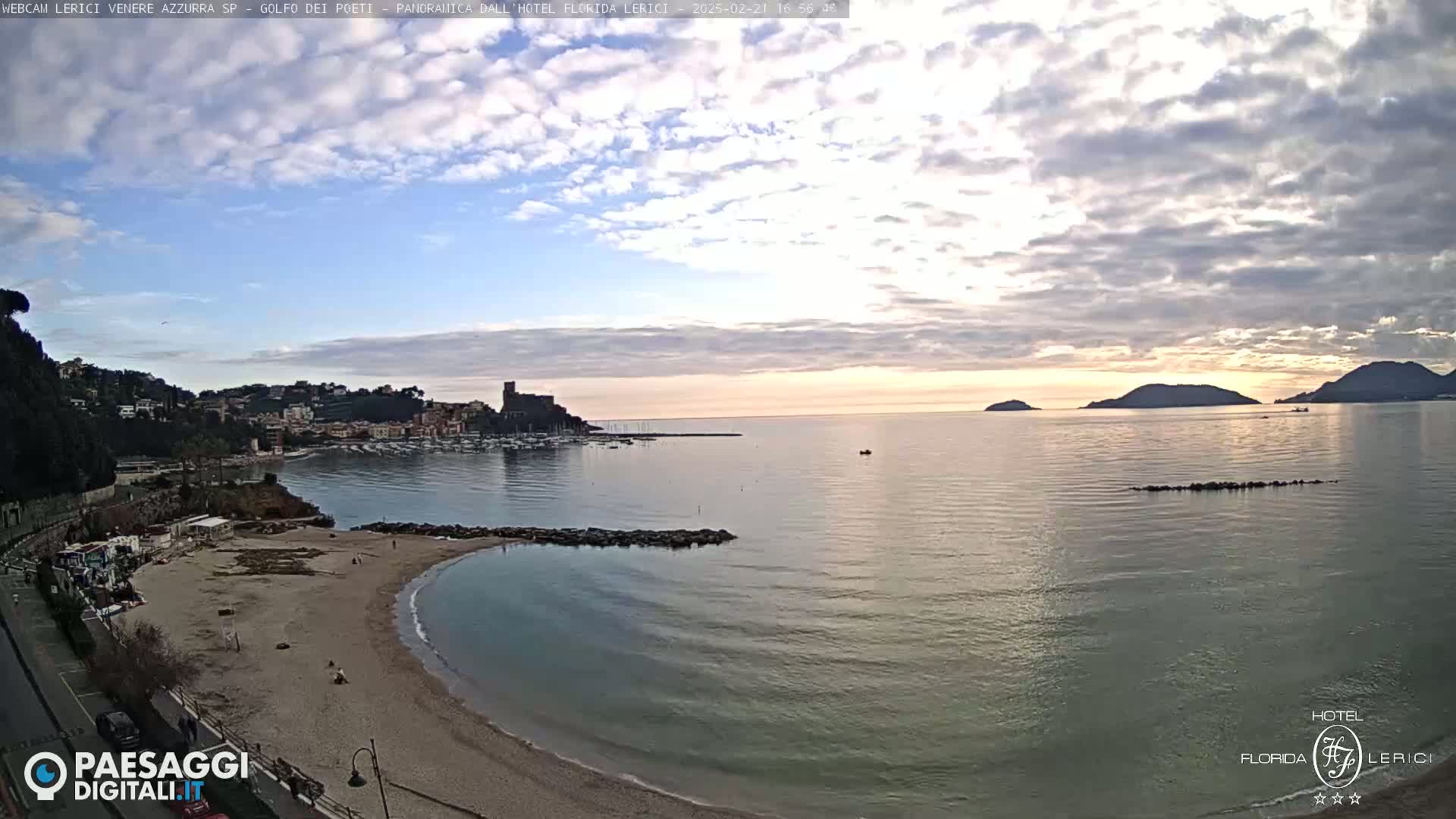 A mostly cloudy, sunset scene overlooking a calm bay with a sandy beach, a town nestled on a hillside, and distant mountains.