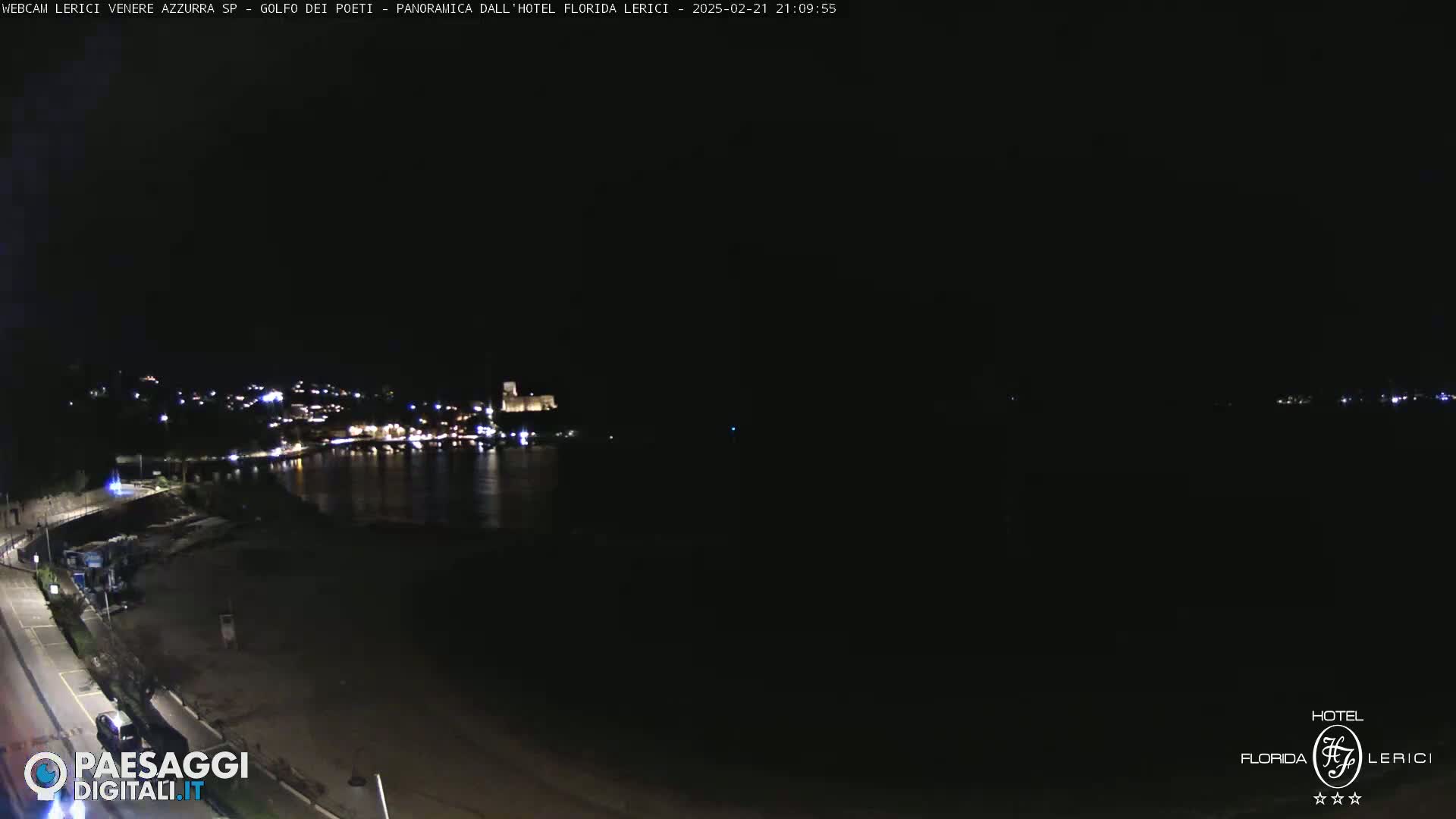 A nighttime coastal scene shows a dark beach, calm water, and a dimly lit town with a castle in the background under a clear, dark sky.