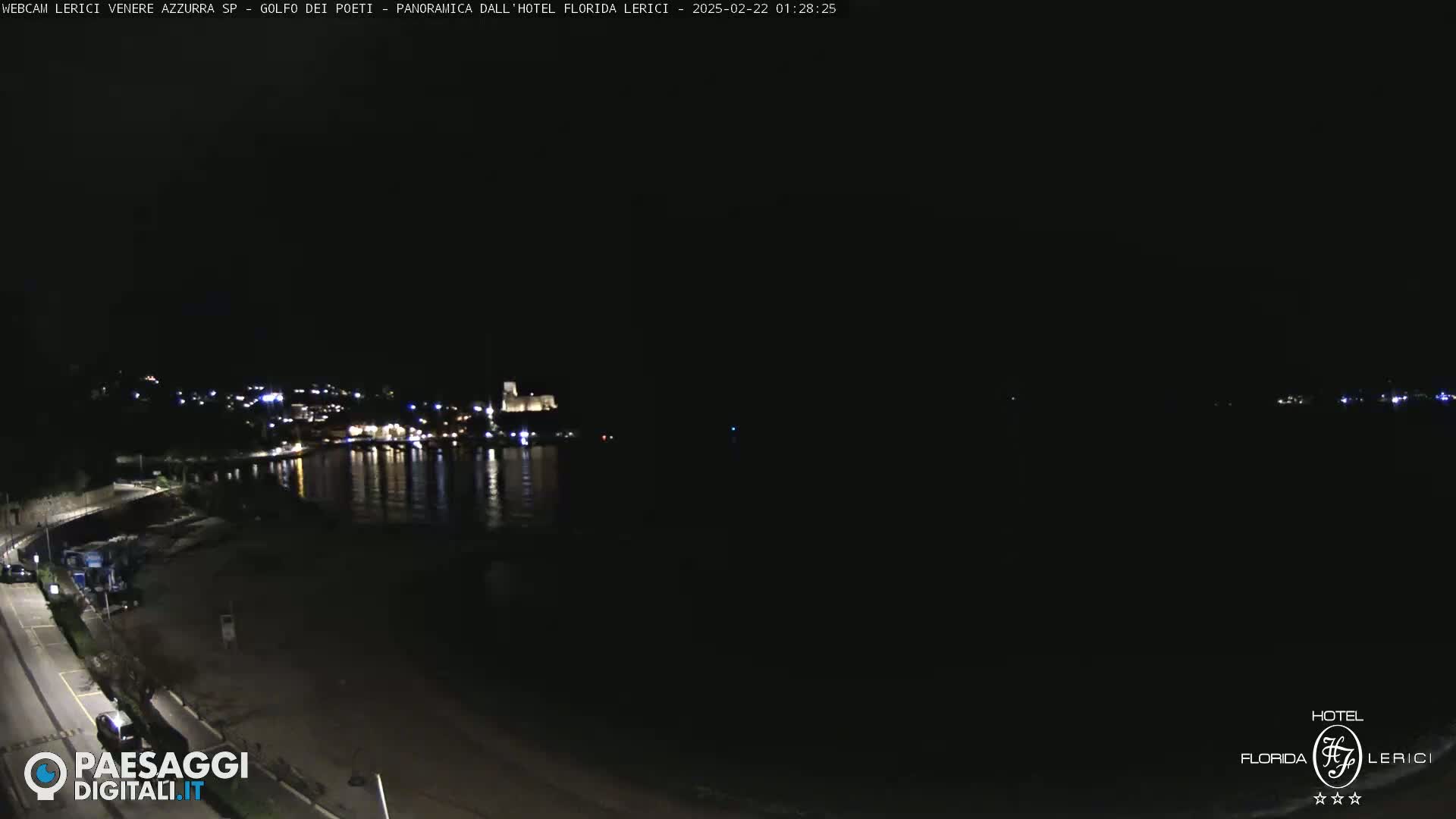A nighttime view of a dark beach and calm water reflecting lights from a town across the water.
