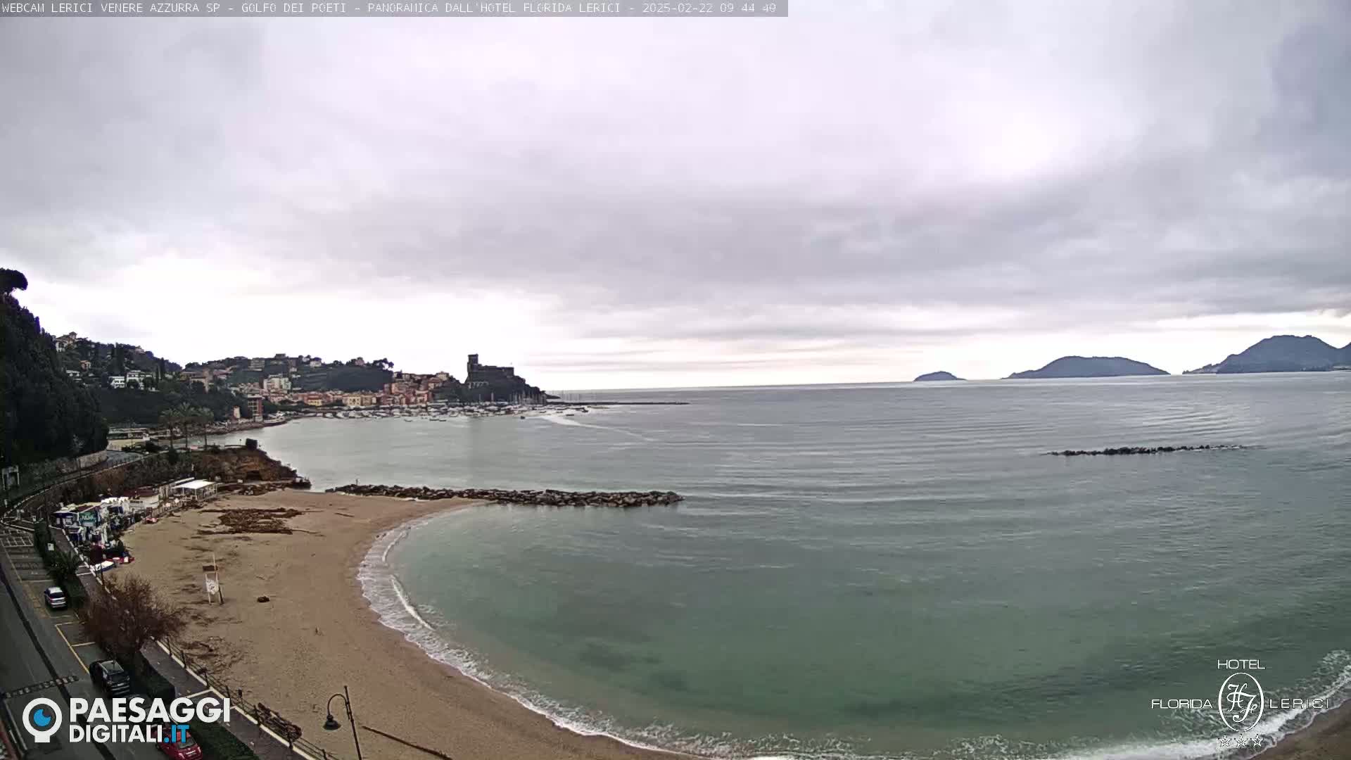 A sandy beach curves into a calm sea under an overcast sky, with a town and hills visible in the distance.