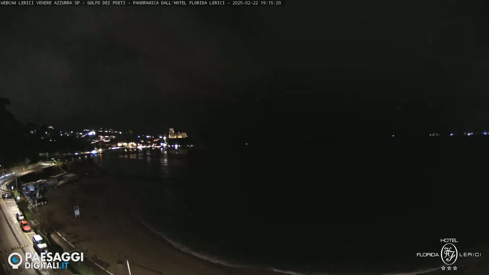 A dark nighttime coastal scene shows a town's lights across a body of water, with a mostly empty beach and a road with a few parked cars in the foreground.