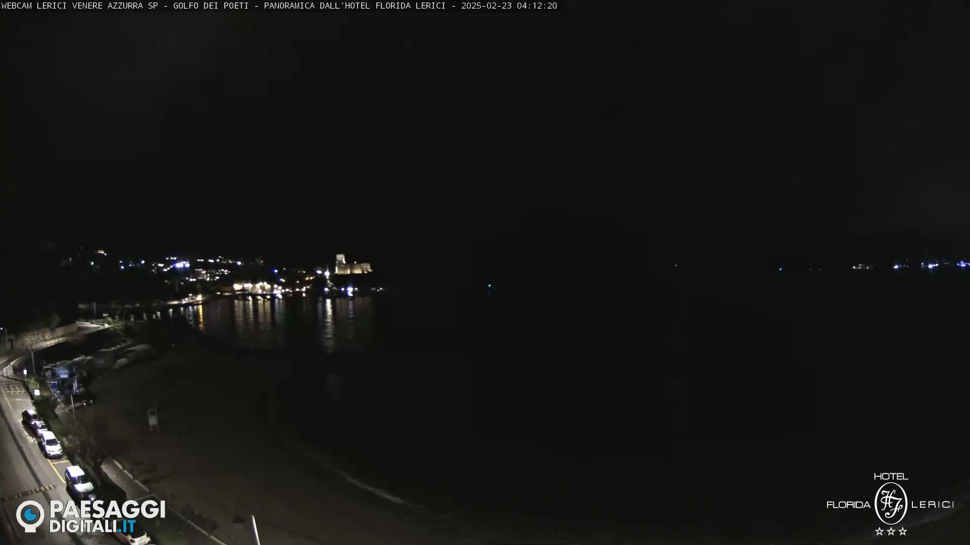 A nighttime, seaside view shows a dark coastline with a few lights in the distance, and a few vehicles parked along a road in the foreground.