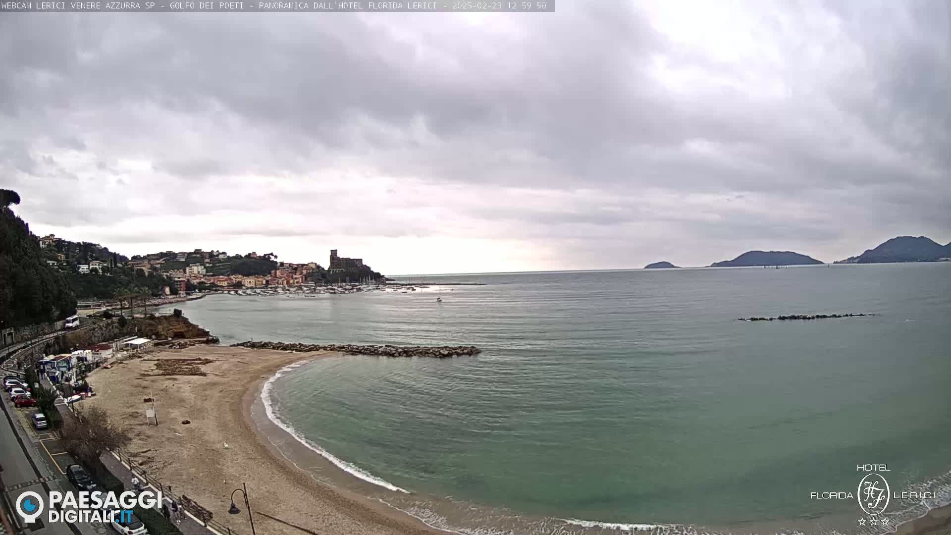 A sandy beach curves into a calm, green-blue sea under an overcast sky, with a town and hills visible in the background.