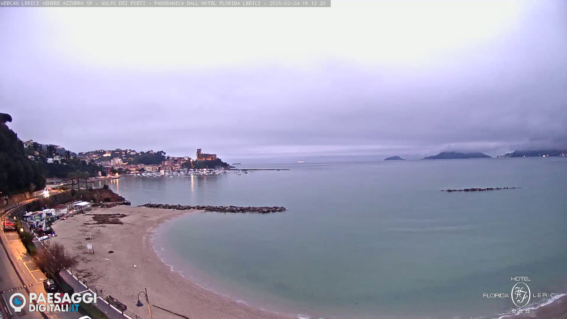 A mostly cloudy twilight scene shows a calm bay with a sandy beach, a small town with lights, and distant hills.