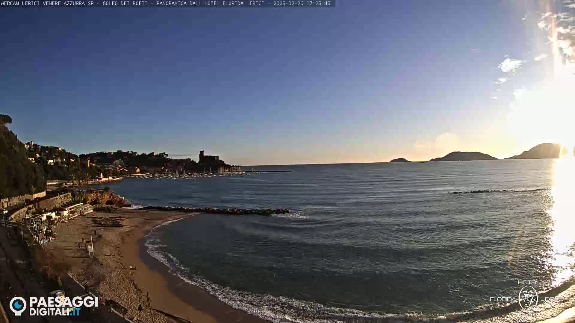 A sandy beach curves into a calm sea under a clear, bright sky, with a town and distant hills visible in the background.