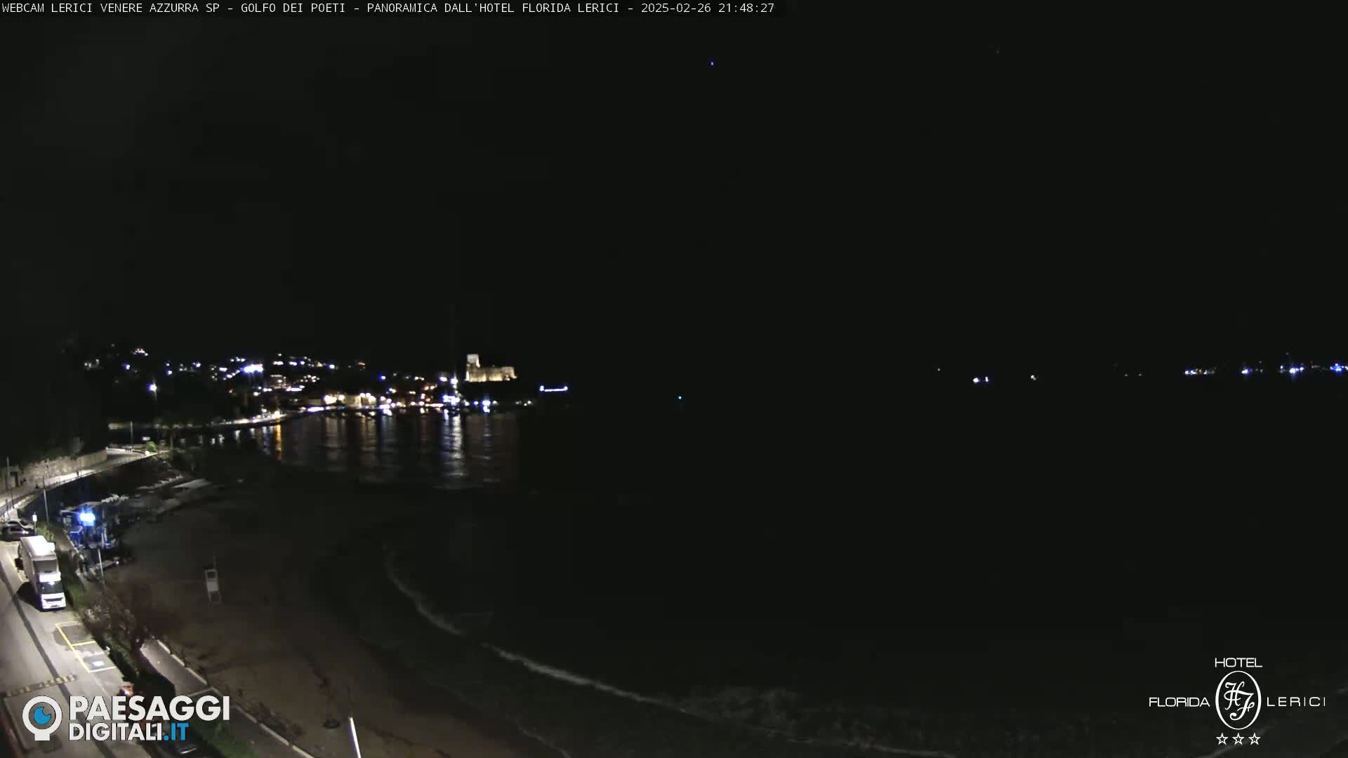 A nighttime view of a dark, sandy beach and calm ocean with a town's lights visible across the water.