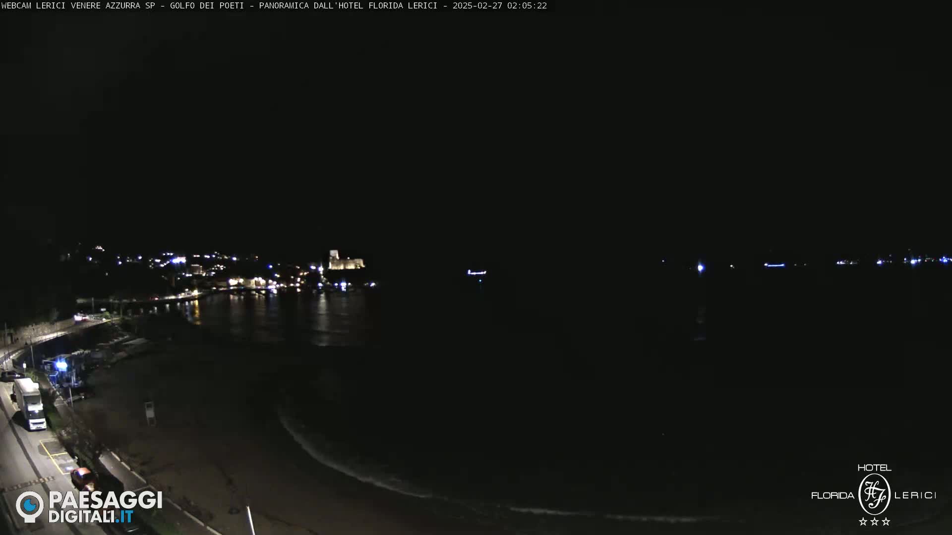 A nighttime view of a dark coastline with a town's lights visible across the water, gentle waves lapping at the shore.