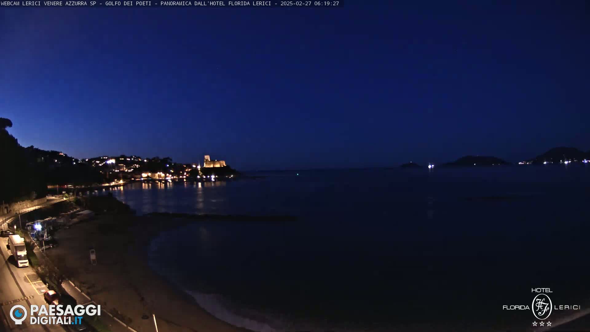 A nighttime coastal scene shows a dark blue sea, a beach, a town with lights, and a hilltop castle, under a clear, dark blue sky.