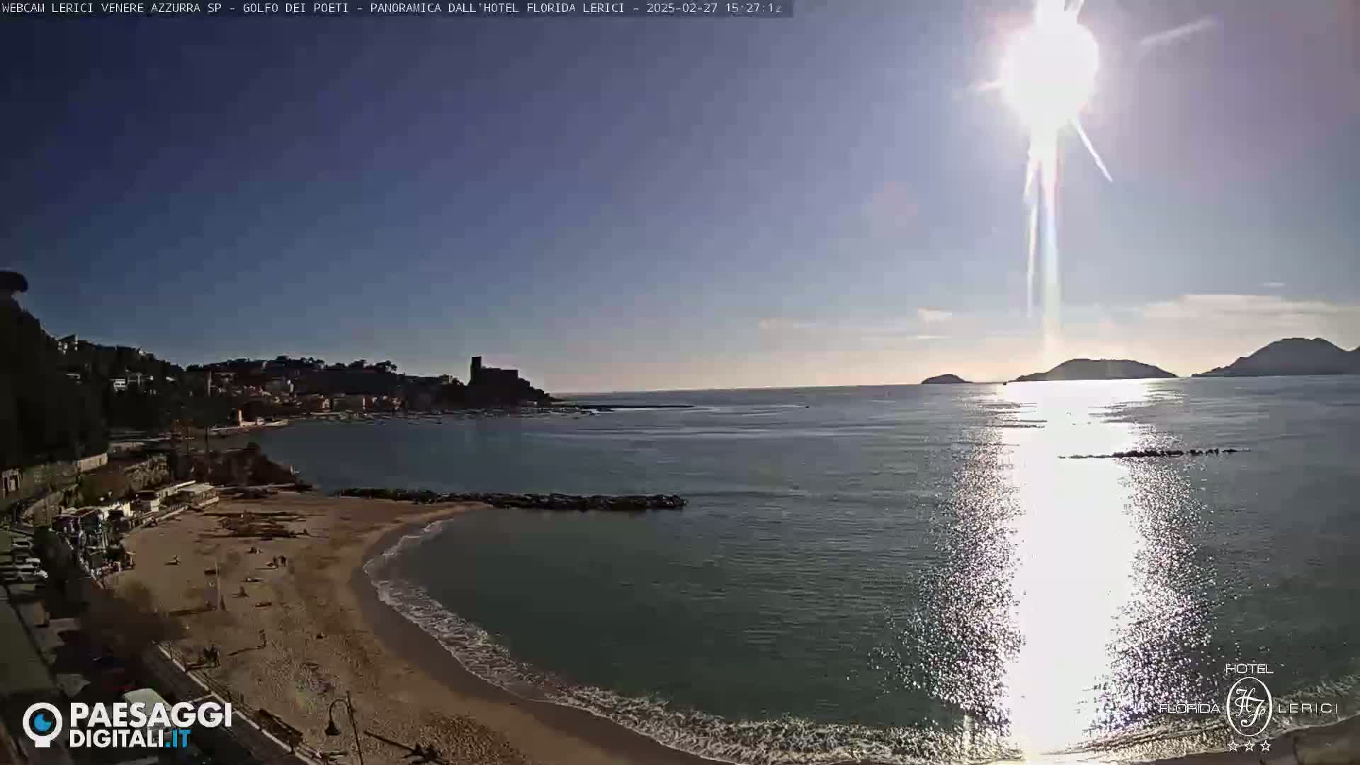 A sandy beach and calm ocean are visible under a bright, sunny sky, with hills and a town in the background.