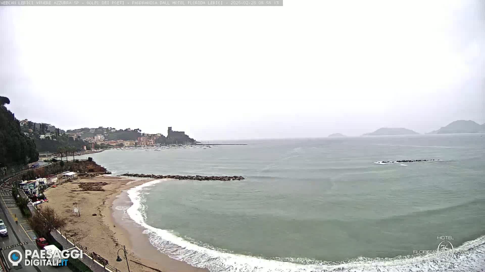 A beach with moderate waves under an overcast sky, overlooking a calm bay with a town and distant hills.