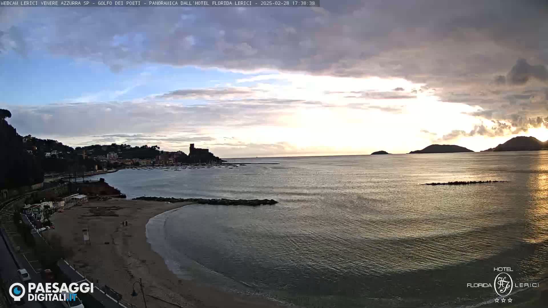 A calm sea at sunset, with a sandy beach, coastal town, and distant hills under a partly cloudy sky.