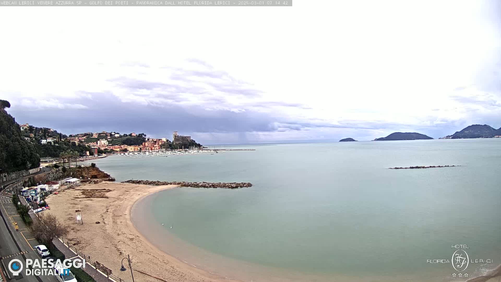 A mostly cloudy day reveals a sandy beach curving into a calm bay, with a town and distant islands visible across the water.