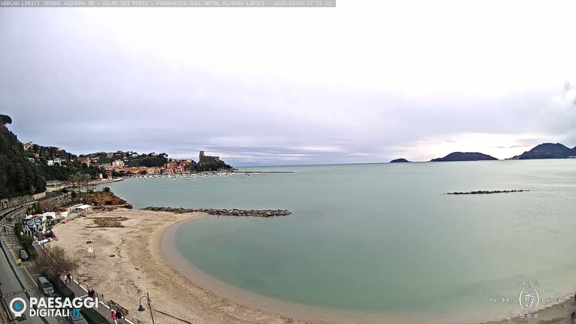 A sandy beach curves into a calm, light-green sea, with a town and distant hills visible under an overcast sky.