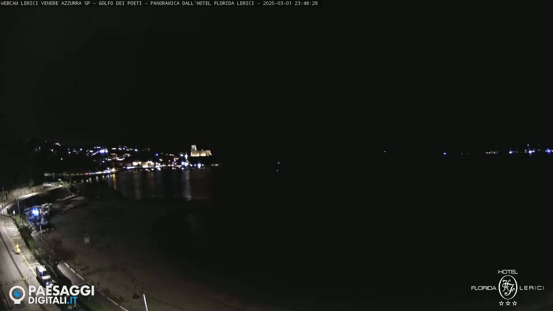 A nighttime coastal view shows a dark beach, a calm body of water, and a town with illuminated buildings across the water.