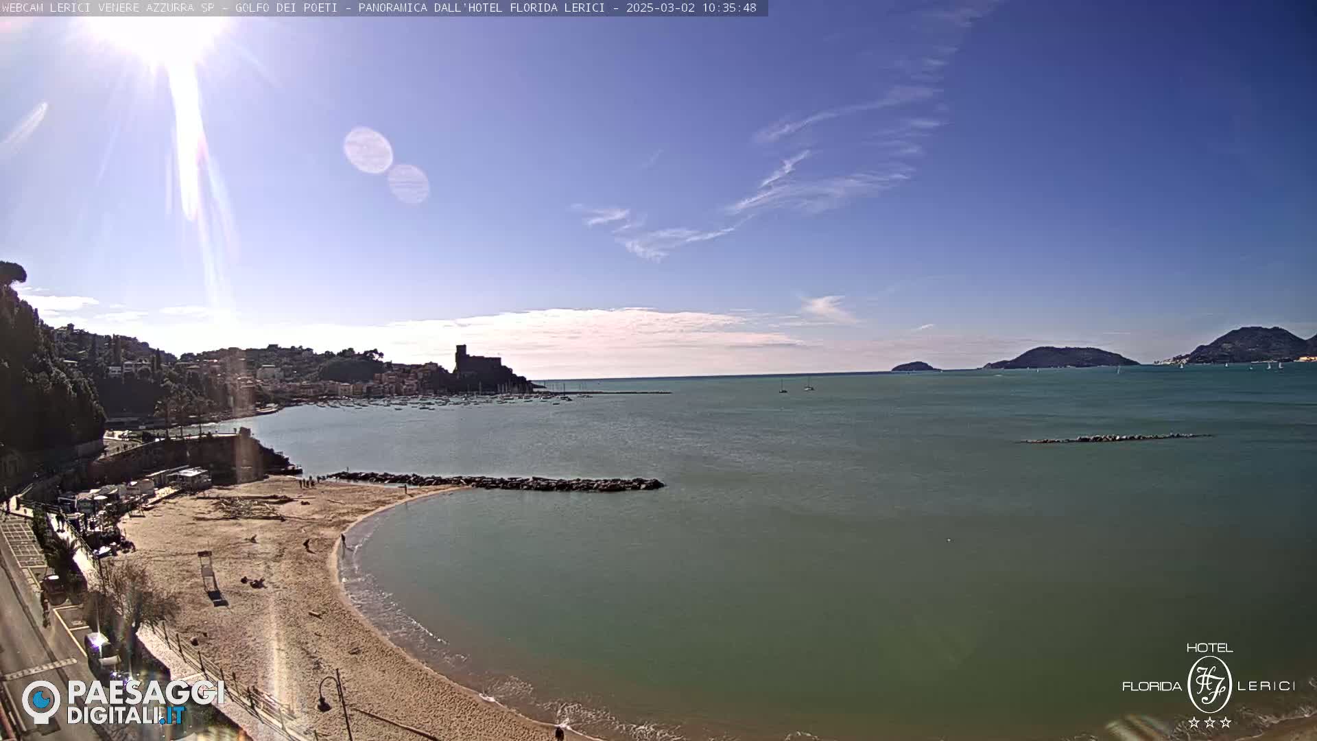 A sandy beach curves into a calm, green-blue sea under a mostly clear, sunny sky, with a town and distant hills visible across the water.