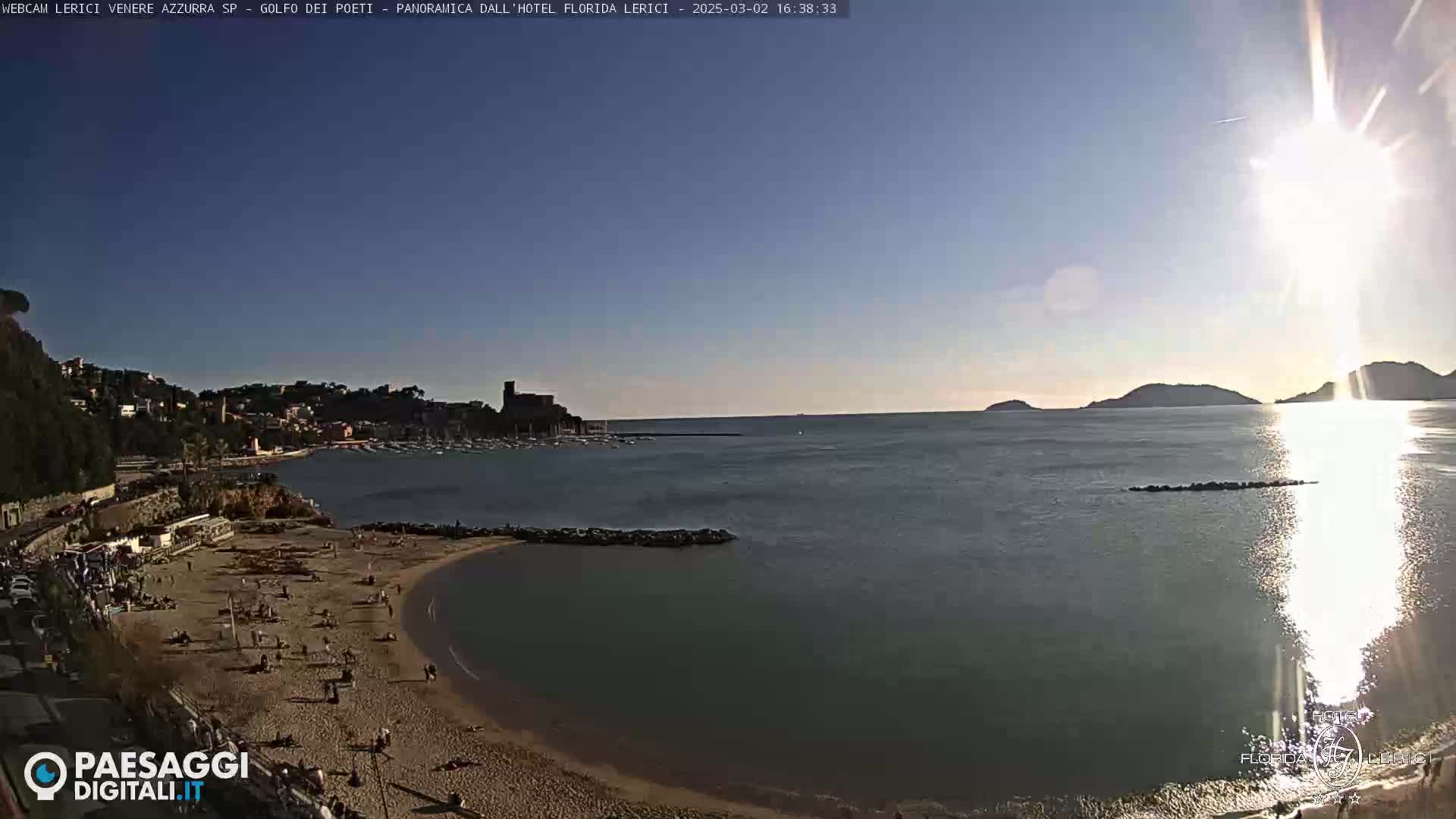 A sunny day at a sandy beach with calm water, a small town in the background, and distant hills.
