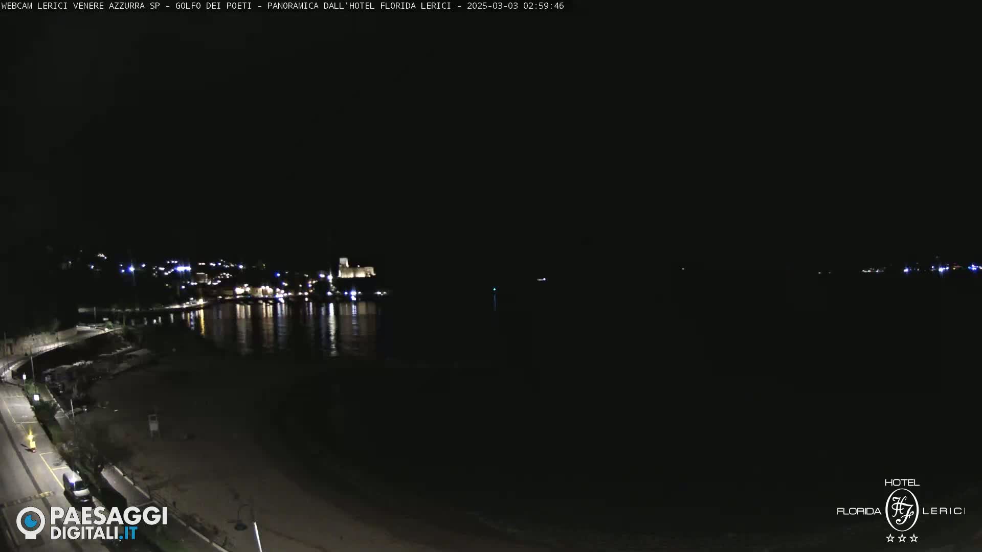 A nighttime, dark view of a beach and calm water reflecting the lights of a town across the water.