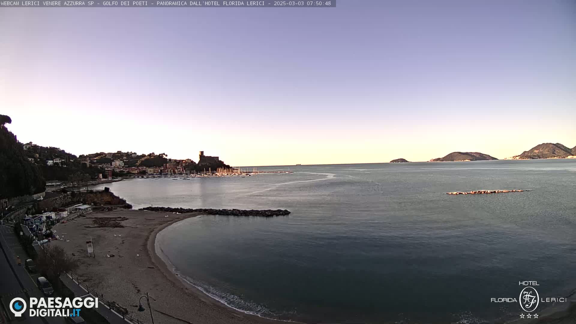 A calm bay with a sandy beach, a small town nestled on a hillside, and boats in the harbor is visible under a clear, twilight sky.