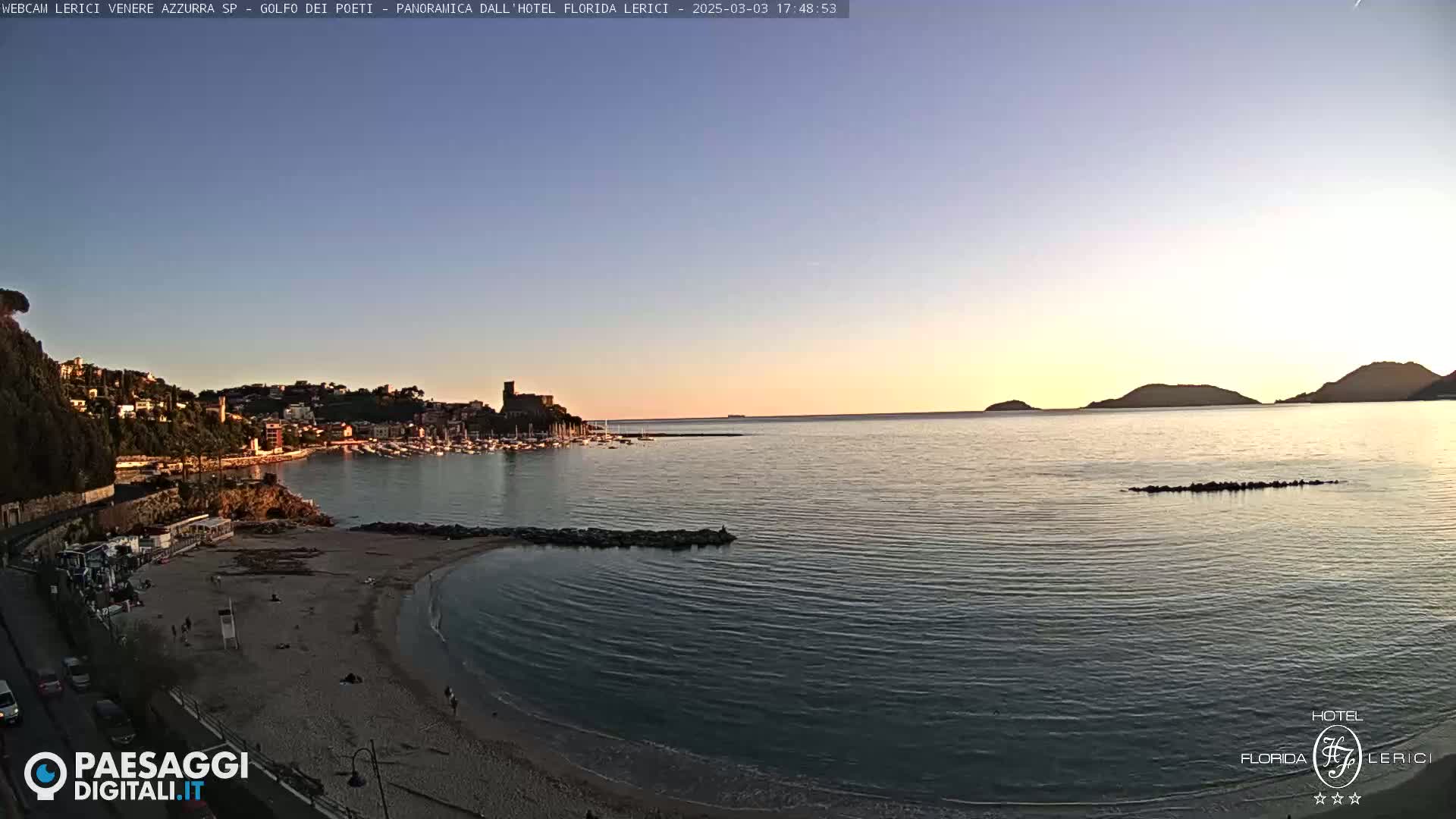 A tranquil beach scene at sunset shows a calm sea, a sandy shore, and a town nestled on a hillside, with distant hills silhouetted against the twilight sky.