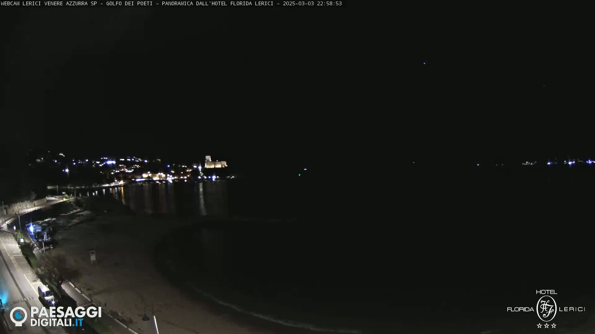 A nighttime coastal scene shows a dark beach, calm water reflecting the lights of a town across the bay, under a clear, dark sky.