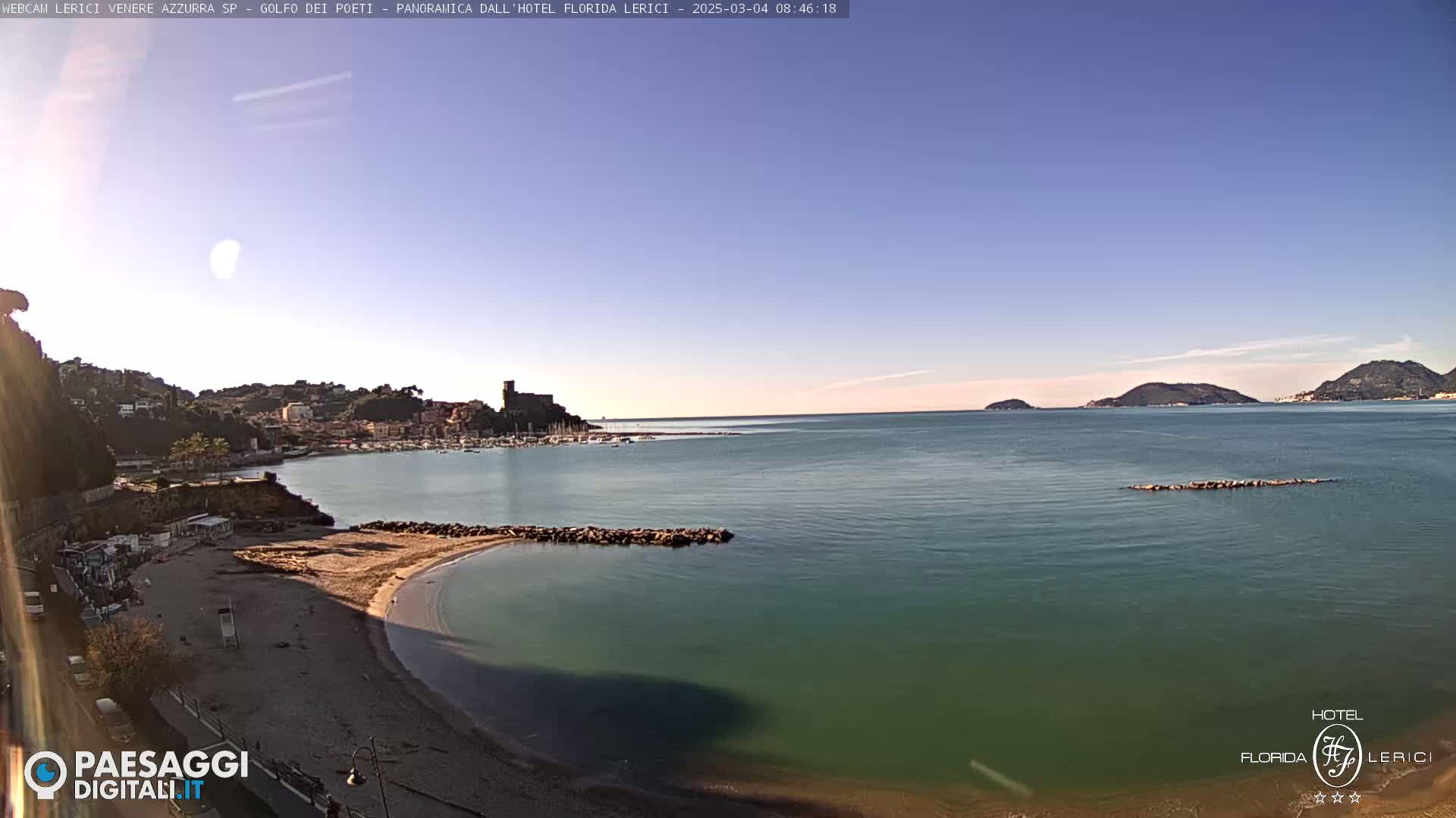 A sandy beach curves into a calm, teal-colored bay, with a town and distant hills visible across the water under a clear, bright sky.