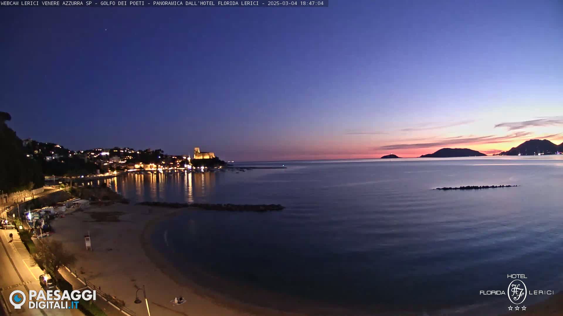 A twilight view of a coastal town with a sandy beach, calm sea, and distant hills under a clear, dusky sky.
