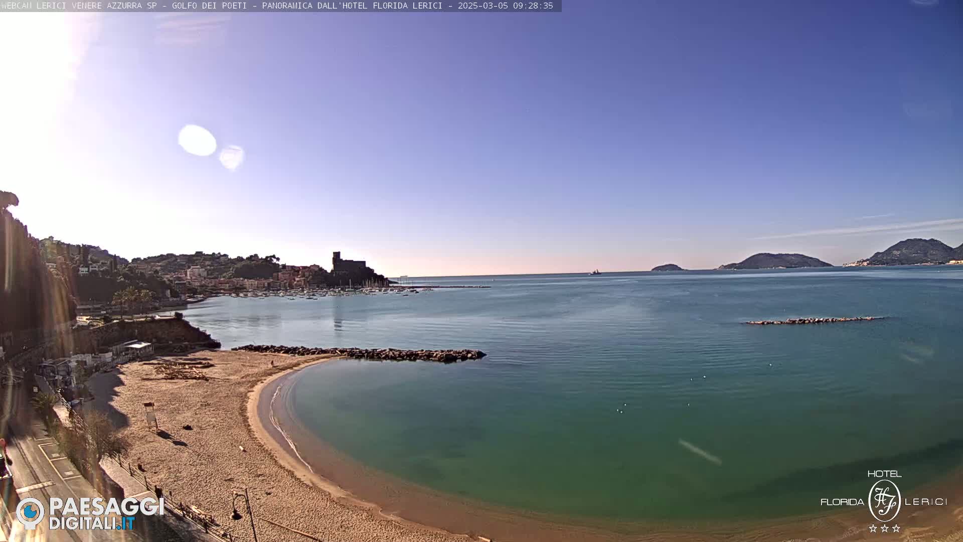 A sandy beach curves into a calm, teal-colored bay, with a town and distant hills visible under a clear blue sky.