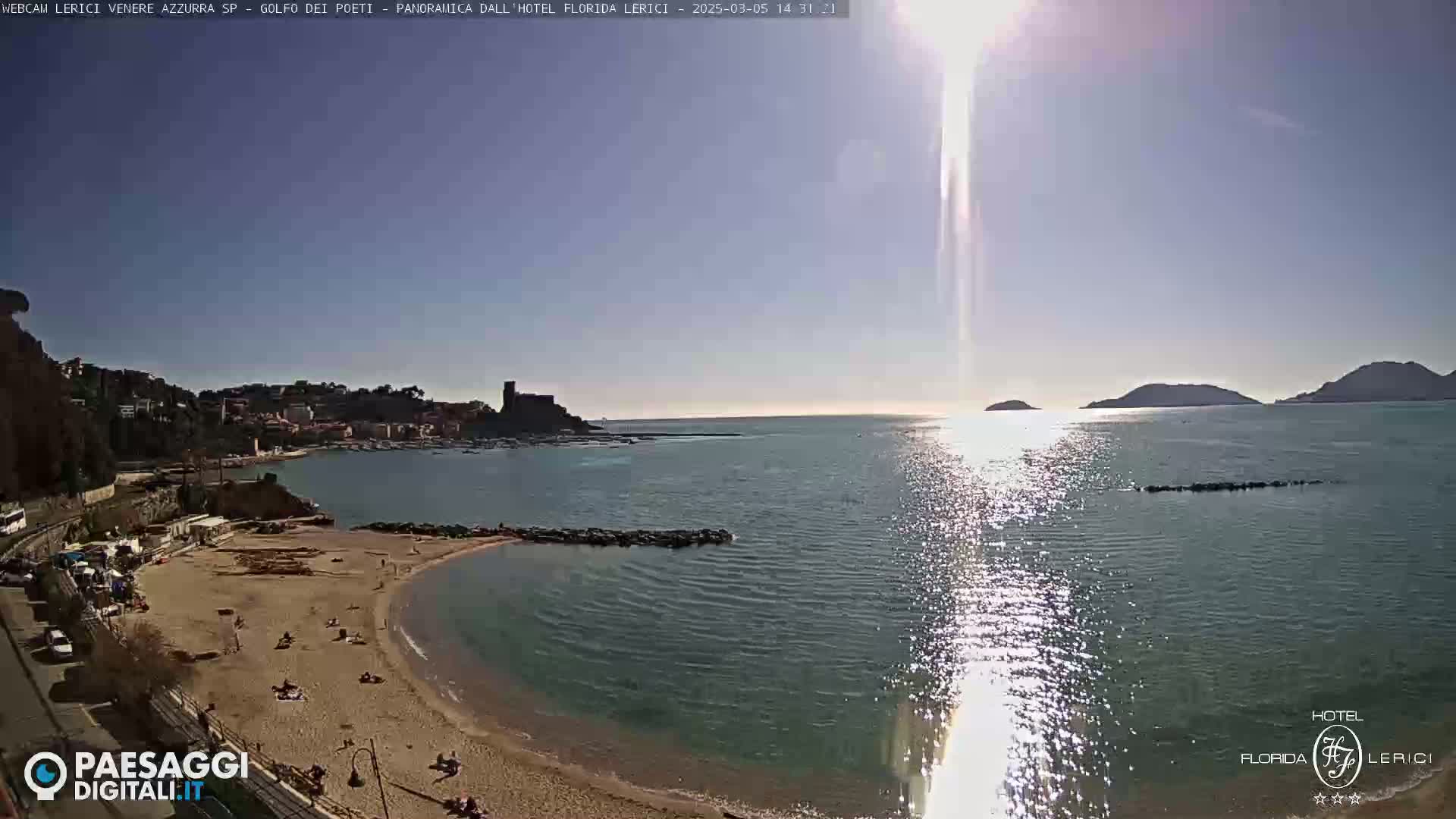 A sandy beach curves into a calm, sunlit bay, with a town and distant hills visible across the water under a clear sky.