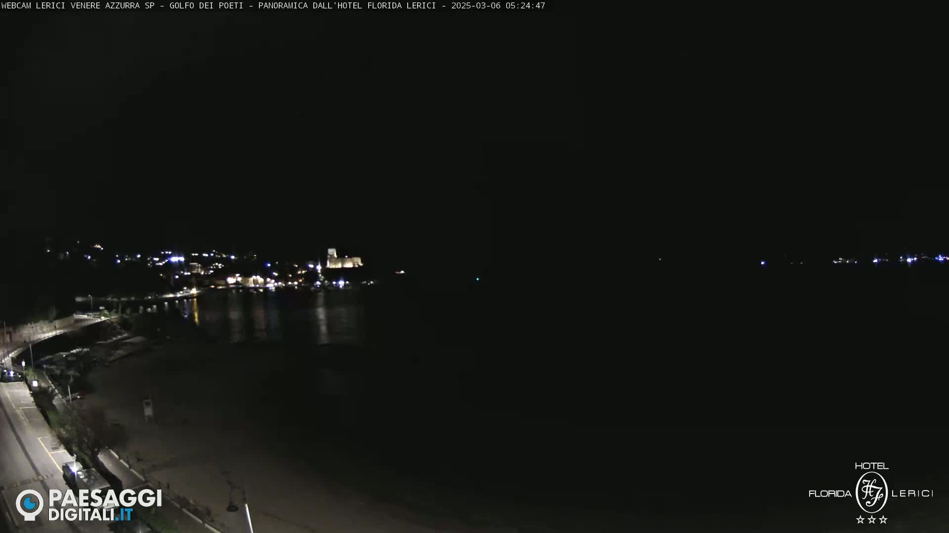 A nighttime, long shot of a dark, calm body of water with a dimly lit town and beach visible in the background.