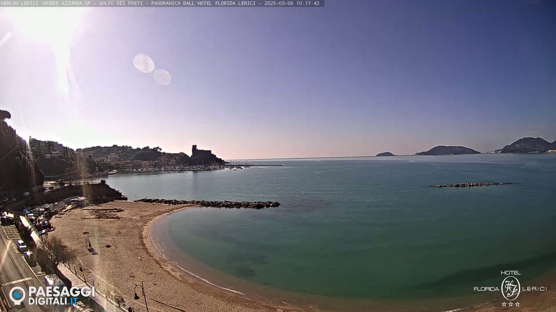 A sandy beach curves into a calm, teal-colored bay, with a town and distant hills visible under a clear, sunny sky.