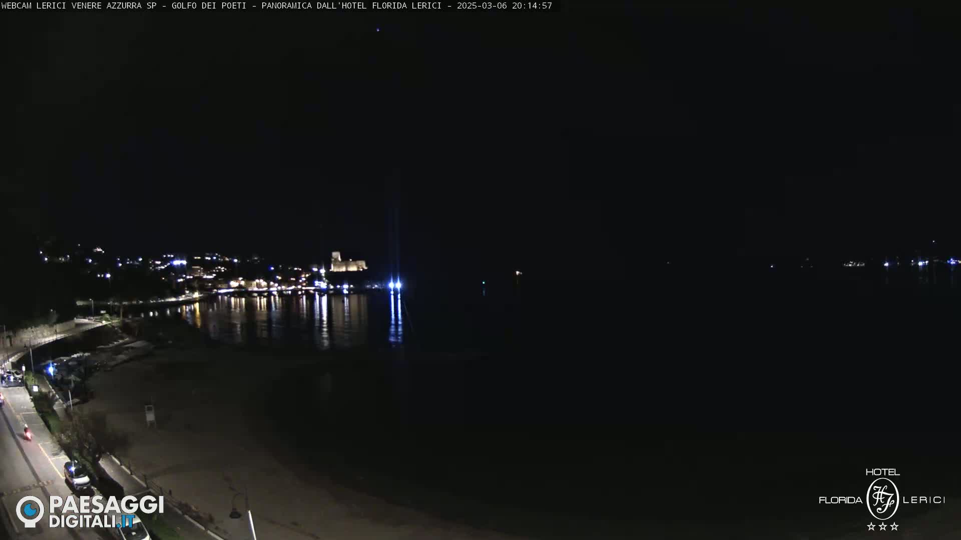 A nighttime view of a dark beach and calm, reflective water with a town's lights visible in the distance.
