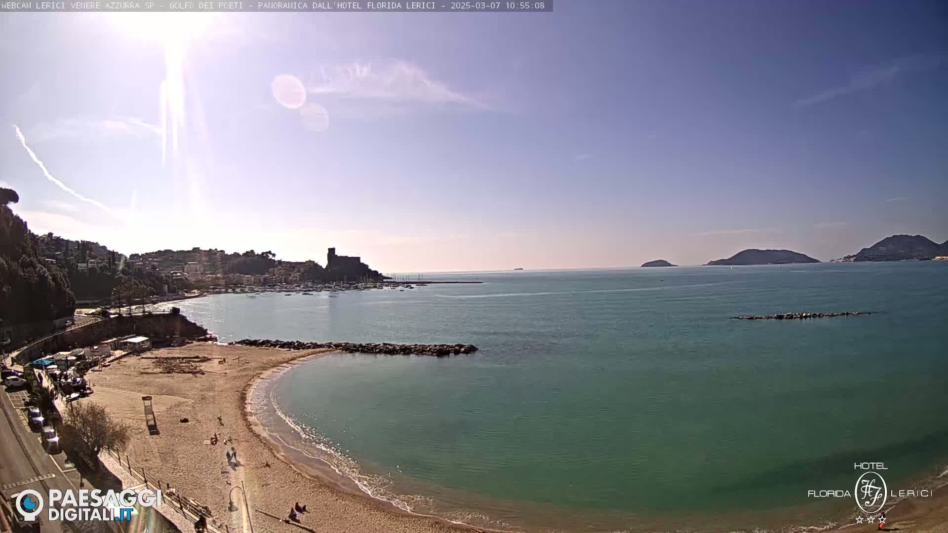 A sunny day reveals a sandy beach curving into a calm, teal sea, backed by a town nestled against green hills and a distant island.