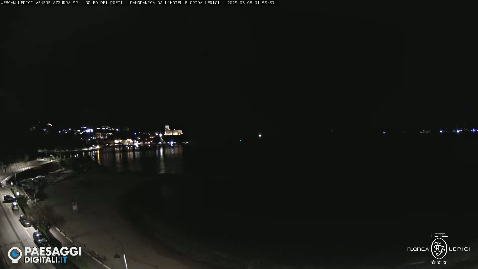 A nighttime, coastal view shows a dark, calm body of water reflecting the lights of a town nestled on a hillside.