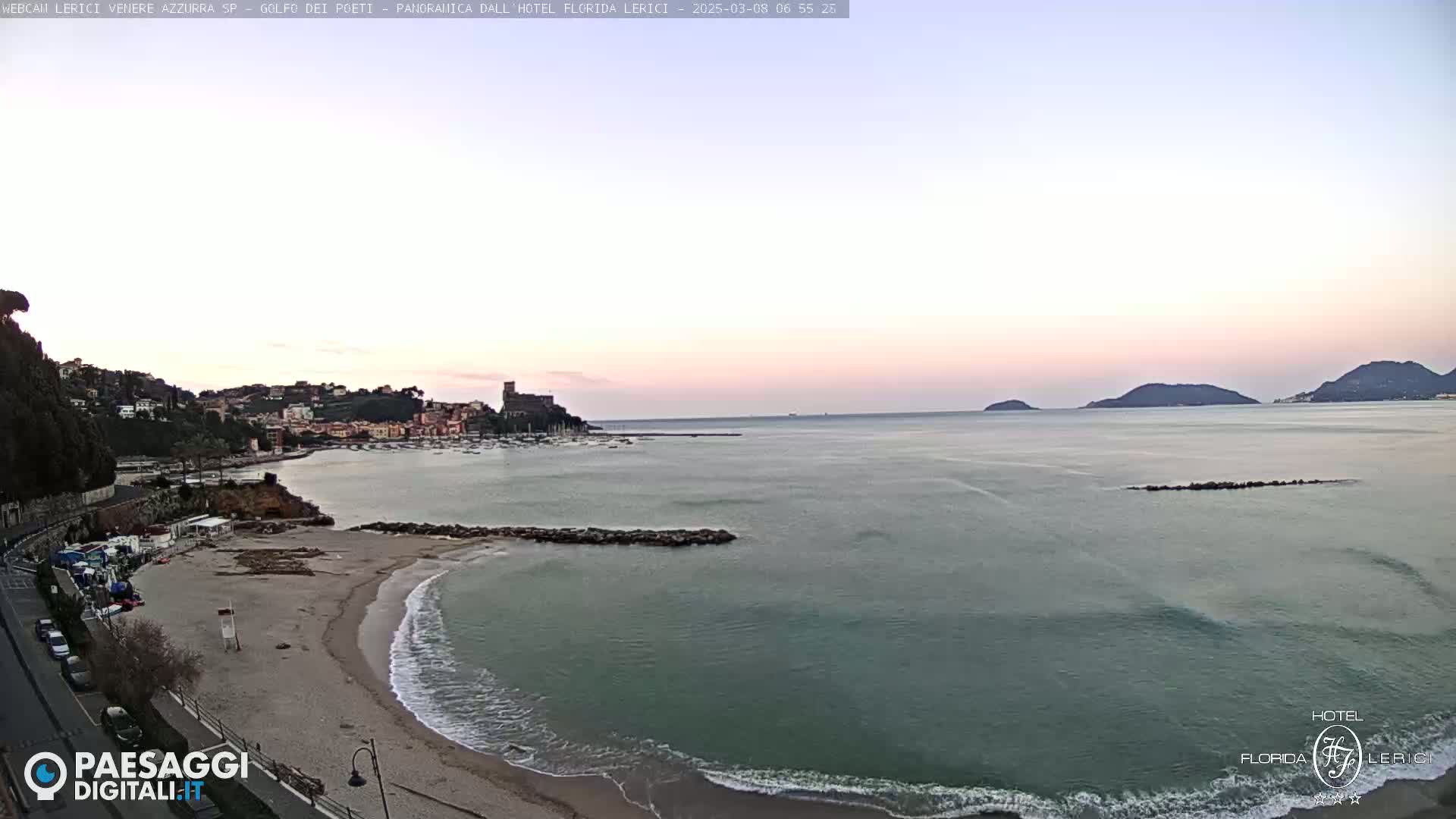 A calm, pale-pink sunrise illuminates a sandy beach, gentle waves lapping the shore, with a town and distant hills visible across a tranquil bay.